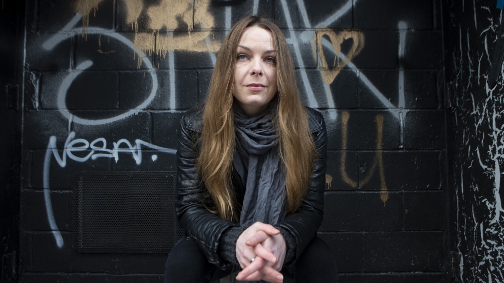 Sonja Mally is photographed near her home in Toronto, on Monday, March 14, 2022. She has long, light brown hair and she is wearing a black jacket. In the background, there are black bricks with silver and gold graffiti. Photo: The Canadian Press/Chris Young.