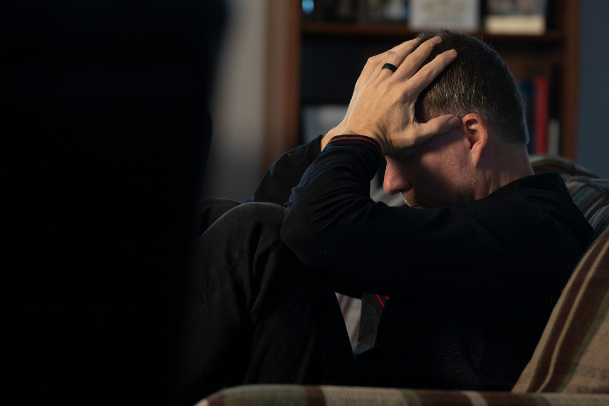 A man sitting on a couch holding his head in his hands. Photo by Malachi Cowie / Unsplash.