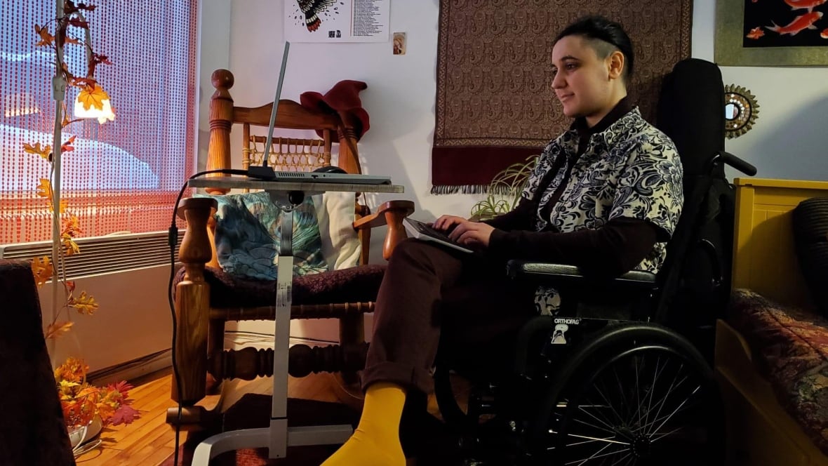 Photo of Nathanael Rafinejad. They are sitting in a wheelchair, in front of a table with a laptop computer. They have short, black hair, and they are typing as they look at the screen.