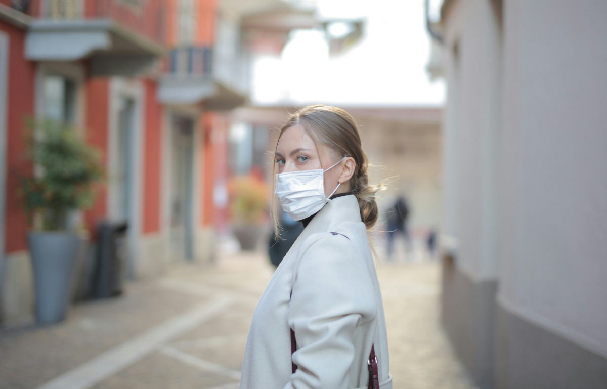 Photo of a woman who is wearing a white coat and a white mask. She has long blond hair. Photo: Bruce Mars / Unsplash.