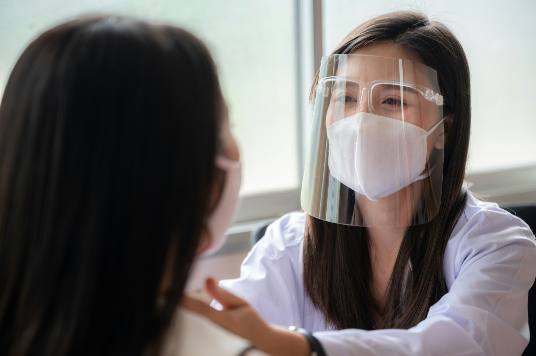 Photo of a doctor (right) who is examining a patient (left). The doctor is wearing a white respirator, and she has long dark hair.