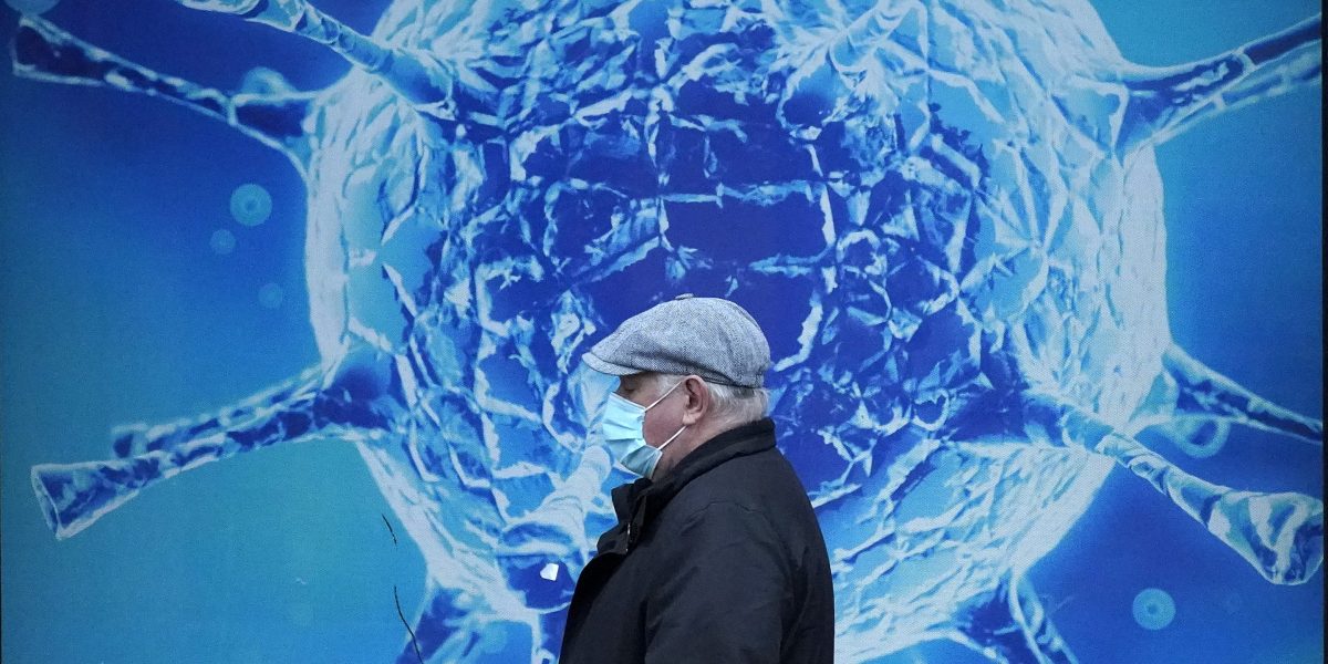 Au premier plan, il y a une personne avec un masque médical bleu et une veste noire. Il y a un fond bleu, avec une image agrandie d’une particule du virus SRAS-CoV-2.