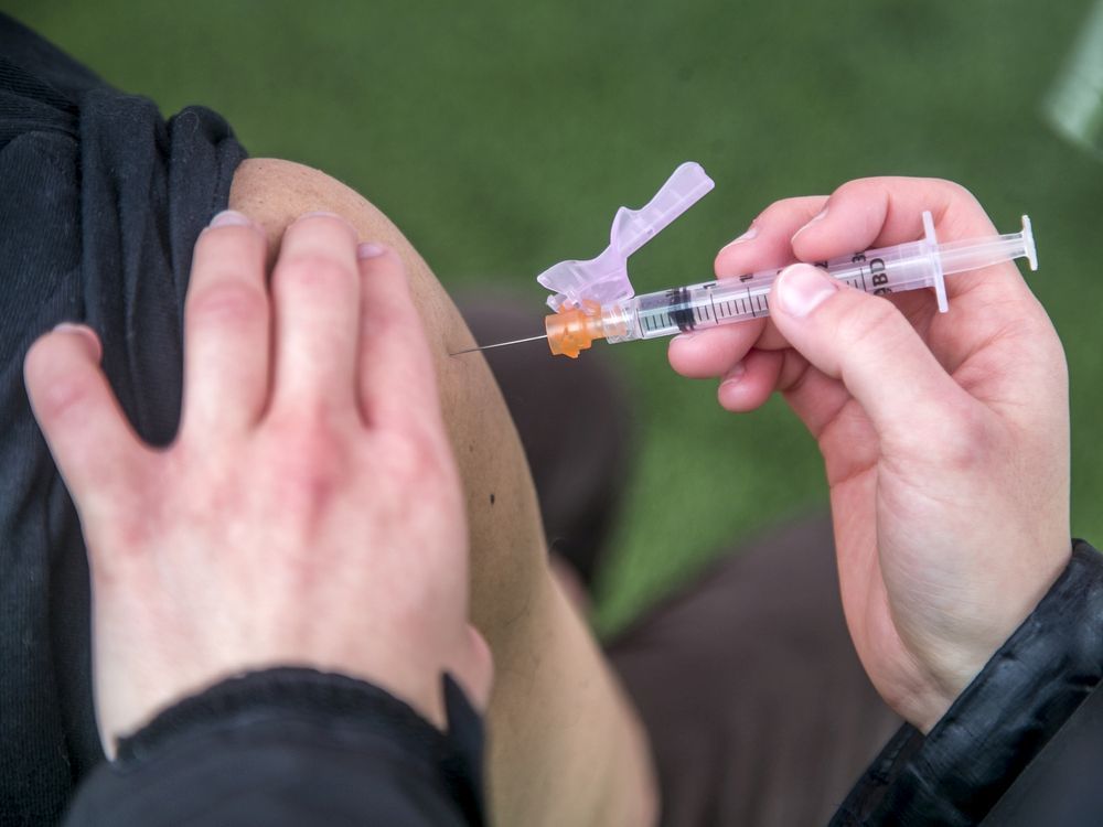 Un travailleur de la santé administre une dose de vaccin à un patient. Le travailleur de la santé tient l’aiguille de sa main droite. Ils portent des vêtements noirs, et il y a un fond vert.