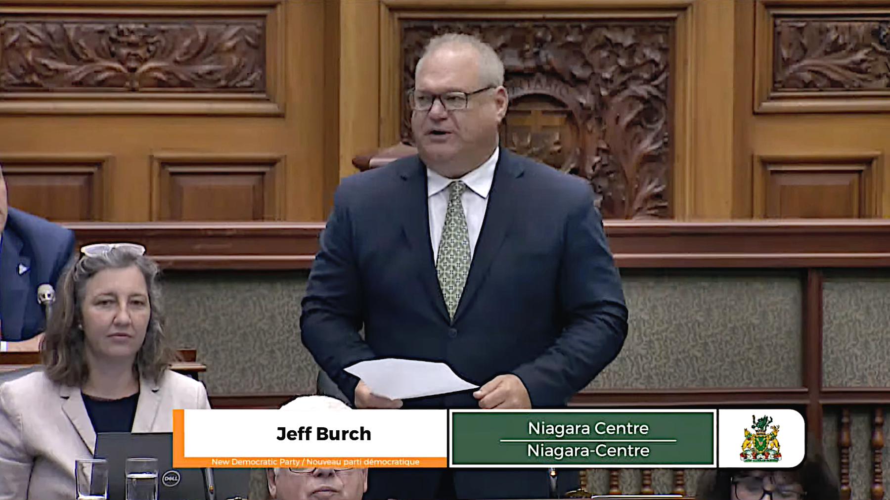 Capture d’écran de la diffusion de la période de questions, le 6 juin 2024. Jeff Burch, député provincial de Niagara-Centre, a des cheveux blancs et gris, des lunettes, une cravate et un costume bleu marine. En arrière-plan, il y a des panneaux de bois sur les murs avec des sculptures complexes.